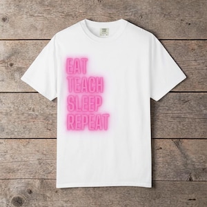 Puede incluir: Camiseta blanca con las palabras "EAT TEACH SLEEP REPEAT" en rosa brillante, apiladas verticalmente. La camiseta tiene cuello redondo y mangas cortas. El fondo es una superficie de madera rústica.