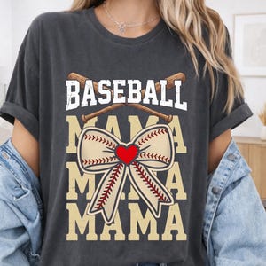 May include: Dark grey t-shirt featuring the text "BASEBALL MAMA MAMA MAMA" in white and tan. The word "BASEBALL" is above crossed baseball bats. A baseball-shaped bow with a red heart is in the center of the design.
