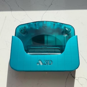 以下が含まれることがあります： 前面に「3D」の文字が入った、ターコイズ色の3Dプリントオブジェクト。 オブジェクトは湾曲した上部と2つのサイドサポートを備えています。 デザインはホルダーまたはケースのようです。