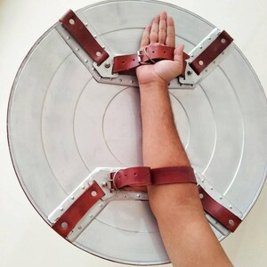 May include: A silver-colored, circular shield with a series of concentric rings. The shield has brown leather straps and metal brackets for securing the shield to the arm. The shield is a replica of a superhero's shield.