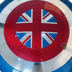 May include: A round shield with a red center featuring a Union Jack design, surrounded by a silver band and a blue outer rim. A small emblem is on the lower right. The shield is a prop or collectible.