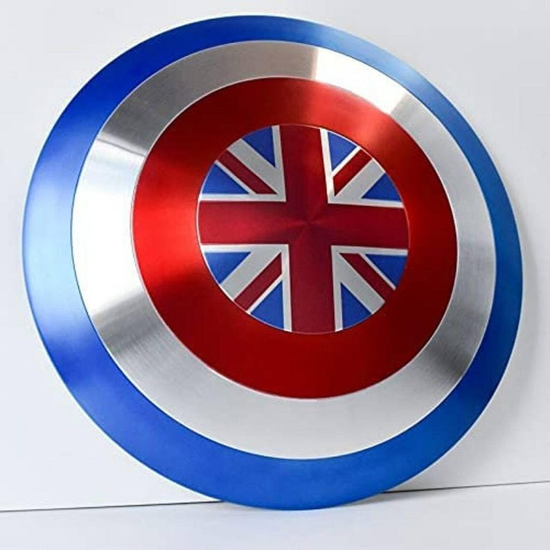 May include: A round shield with a blue, silver, and red color scheme. The center features a red circle with a Union Jack design. The outer edge is blue, with a silver band in the middle. A patriotic themed item.