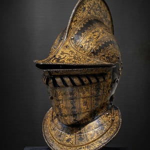 Peut inclure: Casque ancien orné d'un motif floral et géométrique doré sur fond noir. Il est doté d'une visière, d'une protection de cou et d'une crête. Des rivets et une bordure tressée ajoutent des détails. Le casque est présenté sur une surface sombre.