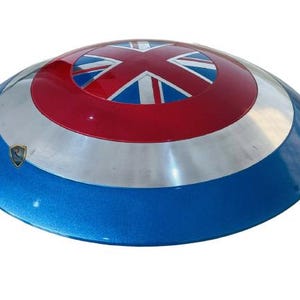 May include: A round shield with a blue, silver, and red color scheme. The shield features a Union Jack design in the center, with a silver band and a blue outer rim. A small emblem is visible on the shield's surface.