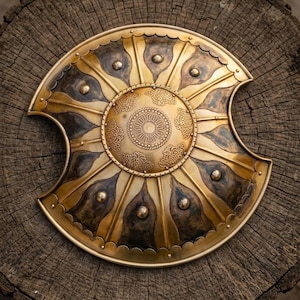 May include: A decorative, antique-style shield with a gold and bronze finish. The shield features a central circular design with intricate patterns and is adorned with rivets. The shield is placed on a weathered wooden surface.