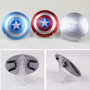 K&ouml;nnte beinhalten: Drei Captain America Schilde in Blau, Rot und Silber, jeweils mit einem Sternemblem. Die Schilde sind rund und werden auf transparenten St&auml;ndern pr&auml;sentiert, die das ikonische Design zeigen.