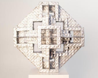 May include: A geometric, silver-colored sculpture with a textured surface, displayed on a white pedestal. The artwork features a symmetrical design with square and rectangular cutouts, creating an open, lattice-like structure. The sculpture is a modern art piece.