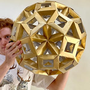 May include: A large, geometric, golden sphere with a complex, open design. The sphere is held by a person wearing a white t-shirt with a deer graphic. The sphere's structure is made of folded, metallic material.