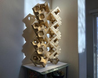May include: A gold-colored, geometric sculpture with a complex, openwork design. The sculpture is composed of interconnected, repeating shapes, creating a visually striking, three-dimensional form. It stands on a glass-topped table, illuminated by natural light.