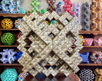 May include: A large, geometric, gold-colored woven art piece is displayed on a small, round, brown table. The artwork is the focal point, with a complex pattern of intersecting lines. Various colorful, geometric sculptures are on shelves in the background.