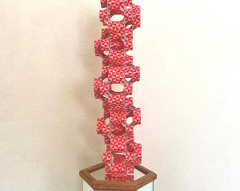 May include: A tall, geometric sculpture composed of stacked, red and white patterned modules. The sculpture is displayed on a mirrored, hexagonal pedestal with a wooden frame. The design features repeating triangular patterns.