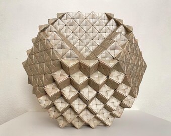 May include: A geometric, spherical sculpture with a woven texture. The piece is constructed from small, square, light-colored woven elements, creating a complex, multi-faceted design. The overall shape is round with a textured surface.