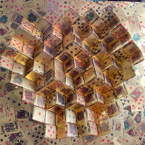 May include: A three-dimensional geometric sculpture constructed from numerous folded playing cards with gold-toned backs. The structure features a repeating pattern of cubes and triangular prisms, showcasing various card suits and face card designs.