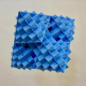 May include: A geometric sculpture composed of light blue, folded paper modules. The artwork has a three-dimensional, textured surface with a repeating diamond pattern. The overall shape is a square with protruding points.