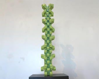 May include: A tall, abstract sculpture with a geometric design. The sculpture is primarily green with a textured surface and a repeating pattern. It stands on a dark, rectangular base against a white wall.