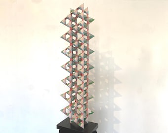 May include: A tall, geometric sculpture with a repeating triangular pattern. The sculpture is composed of interconnected, open-faced pyramids in shades of red, green, and white. It stands on a small, black pedestal against a white wall.
