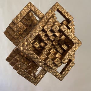 May include: A geometric, gold-colored, woven art piece. The sculpture is composed of multiple square frames, creating an intricate, three-dimensional design. The piece is suspended, showcasing its complex structure and texture.