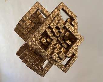 May include: A geometric, gold-colored, woven art piece. The sculpture is composed of multiple square frames, creating an intricate, three-dimensional design. The piece is suspended, showcasing its complex structure and texture.