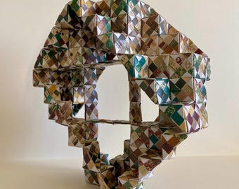May include: A geometric sculpture made of folded paper, featuring a repeating diamond pattern in shades of brown, green, and cream. The artwork has an open, arch-like structure with a staircase design, creating a three-dimensional effect.