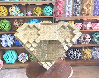 May include: A large, gold-colored geometric origami sculpture, diamond-shaped, with a square opening. The sculpture is on a wooden table, with a wooden shelf in the background filled with colorful origami models.