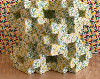 May include: A woven cube-shaped decorative object. The object is constructed from small, woven squares in yellow, green, and white. The background features a woven pattern in red, yellow, and green. The object is sitting on a wooden surface.