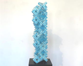 May include: A tall, abstract sculpture composed of stacked, light blue geometric shapes. The sculpture has a repeating pattern of diamond shapes within each cube, and it stands on a black pedestal against a white background.