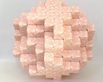 May include: A geometric sculpture composed of numerous small, light pink paper cubes. The cubes are arranged in a spherical shape, creating a textured surface with a woven pattern. The overall form is symmetrical and visually appealing.