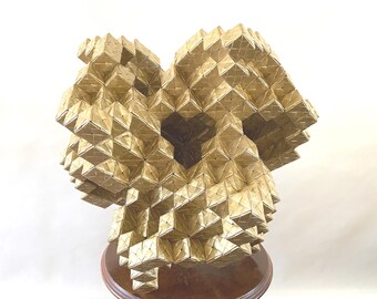 May include: A gold-colored, geometric sculpture with a complex, multi-faceted design. The sculpture is composed of numerous small, square-shaped elements arranged in a three-dimensional form. It sits on a round, wooden base, creating a striking contrast.