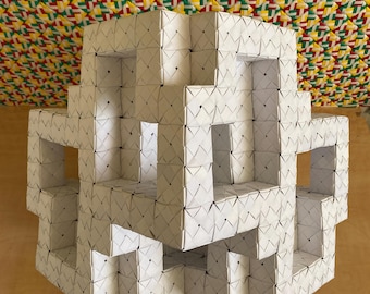 May include: A white, geometric sculpture composed of folded paper modules. The structure has an open, cube-like design with multiple rectangular openings. The background features a colorful woven pattern in red, yellow, green, and white.
