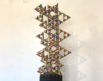 May include: A colorful geometric sculpture with a layered design. The sculpture is composed of interconnected triangular and hexagonal shapes, painted in shades of blue, green, yellow, and red. It stands on a black base against a white wall.