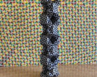 May include: A tall, geometric sculpture with a black and white triangular pattern. The sculpture is set against a colorful background of woven fabric in red, yellow, green, and white. The sculpture is on a wooden surface.