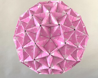 May include: A geometric, spherical paper art piece. The sphere is constructed from pink and white woven paper, creating a complex pattern of triangles and diamond shapes. The paper art is suspended from a thin white string.