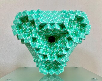 May include: A geometric sculpture in the shape of a stylized head, crafted from folded paper. The sculpture features a repeating pattern of green and white triangles, creating a textured surface. The sculpture has a dark, circular opening in the center.