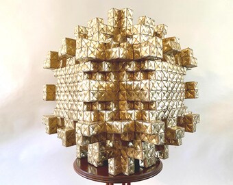 May include: A golden, geometric sculpture composed of numerous small, square-shaped elements. The sculpture has a spherical form, with a textured surface. It is displayed on a dark wooden, round table.