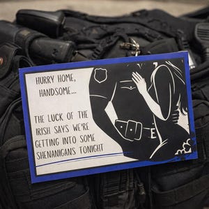 Puede incluir: Una tarjeta de felicitación con borde azul, con una ilustración en blanco y negro de un oficial de policía y una mujer abrazándose. El texto de la tarjeta dice: "Date prisa en casa, guapo... La suerte de los irlandeses dice que vamos a hacer travesuras esta noche."