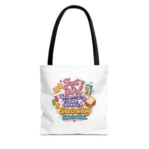 May include: A white tote bag with black handles featuring a colorful graphic print that reads Just a girl who needs her little Snacky snacks. The design includes illustrations of crackers, gummy bears, a juice box, and a sandwich.