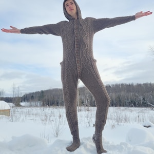 May include: A brown hooded jumpsuit with a cable knit design. The one-piece garment features a button-up front, pockets, and attached feet. The person is standing in the snow with arms outstretched.