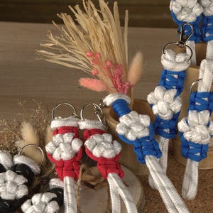 May include: A collection of handmade paracord keychains featuring a woven white paw print design. The keychains are available in blue, red, and black cord accents, each attached to a silver-toned metal lobster clasp and a circular split ring.