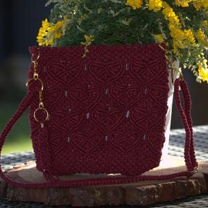 May include: A burgundy macrame crossbody bag with a heart pattern, gold-colored hardware, and a long strap. The bag is displayed on a wooden slab, with a background of yellow flowers and greenery.