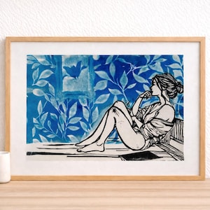 May include: A framed art print featuring a black and white illustration of a person seated, with a thoughtful pose. The background showcases a blue floral pattern. The artwork is in a light wood frame.