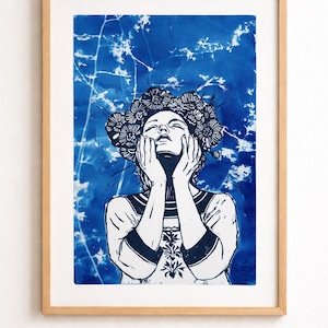 May include: A framed art print featuring a woman with a floral crown, hands on her face, against a blue and white background. The artwork is in a wood frame, creating a striking contrast. The print has a unique, artistic style.