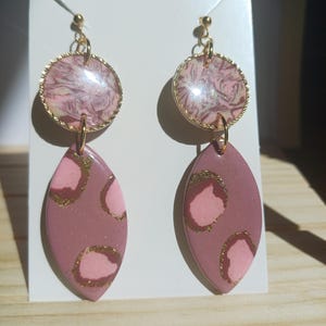 May include: A pair of dangle earrings. The top portion is a gold-toned circle with a pink and white marbled design. The bottom portion is an oval shape in a mauve color with pink and gold accents. The earrings are attached with gold-toned hardware.