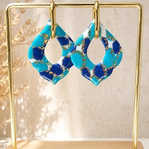 Op de afbeelding: Een paar hangende oorbellen met een ruitvormige hanger met een geschulpte rand. Het oppervlak toont een mozaïekpatroon van witte, turquoise en donkerblauwe segmenten met gouden vlokken, opgehangen aan een goudkleurige metalen ring.