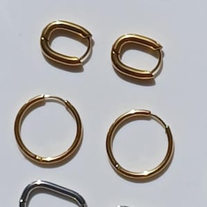 May include: An assortment of gold and silver-toned hoop earrings in various shapes and sizes. The earrings include oval and circular designs, all with a polished finish. The jewelry is displayed on a white surface.
