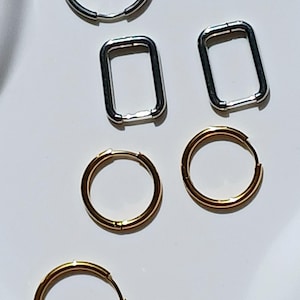 May include: An assortment of hoop earrings in various shapes and colors. The image displays silver and gold-toned earrings, including round and rectangular hoops. The earrings are arranged on a white surface, showcasing their different designs and finishes.