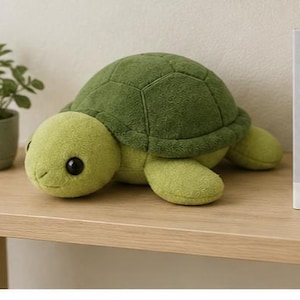 Könnte beinhalten: Ein Schildkröten-Plüschtier mit hellgrünem Kopf und Gliedmaßen sowie einem dunkelgrünen, strukturierten Panzer. Die Schildkröte hat große schwarze Augen und einen gestickten Mund. Sie liegt auf einer Holzoberfläche neben einer Pflanze und einem Buch.