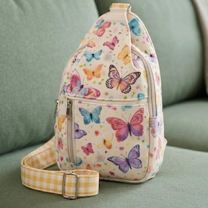 May include: Cream-colored sling bag with a colorful butterfly pattern. The bag features a zippered main compartment, a yellow and white checkered strap, and a small front pocket. The butterflies are in shades of pink, blue, orange, and purple.