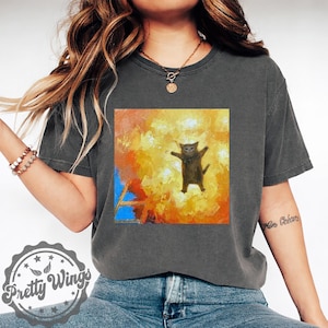 Puede incluir: Camiseta gris oscuro con un gráfico cuadrado de un gato con los brazos extendidos sobre un fondo abstracto y ardiente. La camiseta tiene un corte holgado y está hecha de un material suave. La camiseta tiene el logo de Pretty Wings en la parte inferior izquierda.