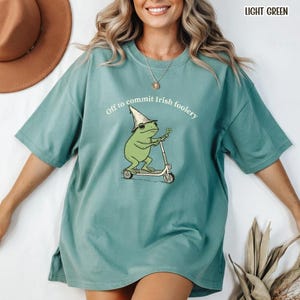 以下が含まれることがあります： 「Off to commit Irish foolery」の文字が入ったライトグリーンのTシャツ。レプラコーンの帽子とサングラスをかけた漫画の蛙がスクーターに乗り、四つ葉のクローバーを持っています。