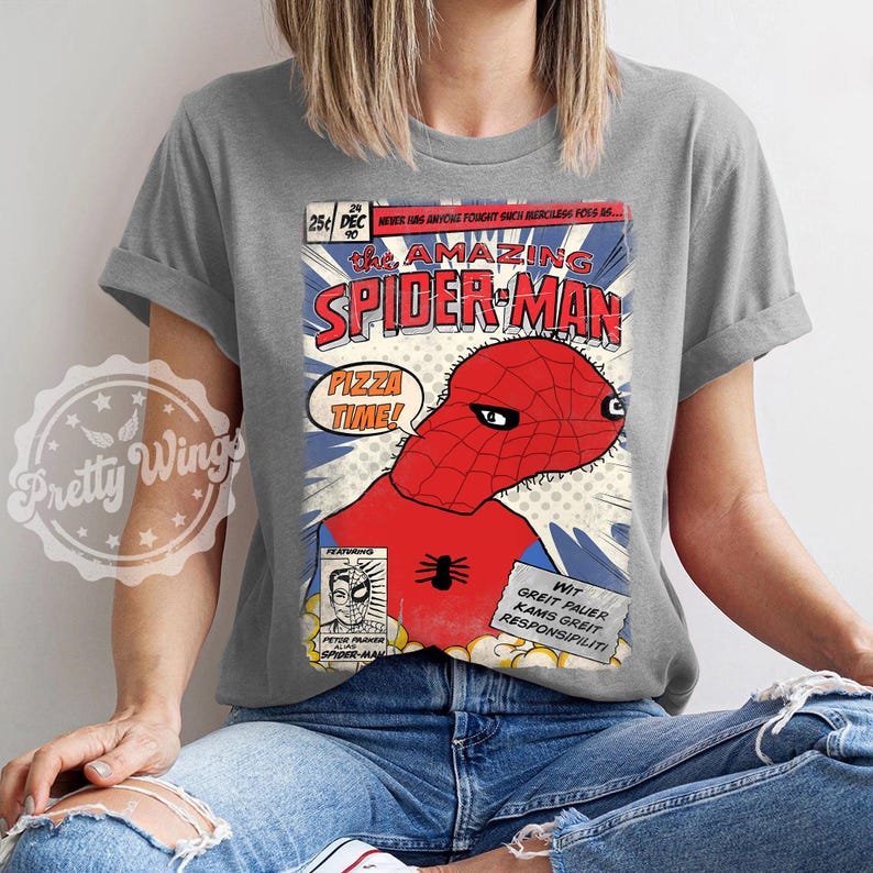 May include: A gray t-shirt featuring a comic book-style illustration of Spider-Man. The design includes the text "The Amazing Spider-Man" and "Pizza Time!" in a speech bubble. The shirt has rolled-up sleeves.